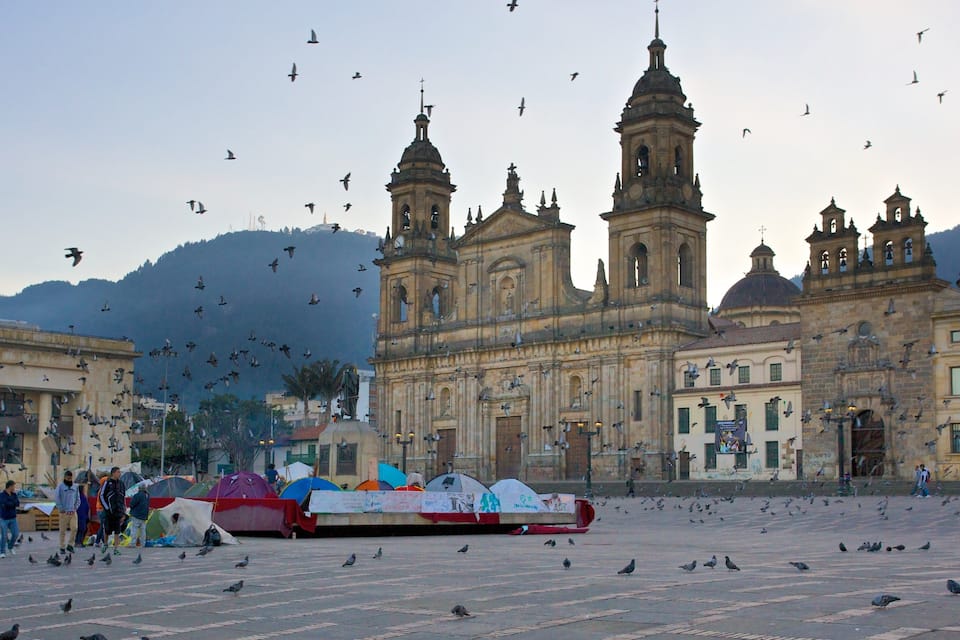 A historic cathedral with multiple towers and domes, surrounded by pigeons and people in a plaza.