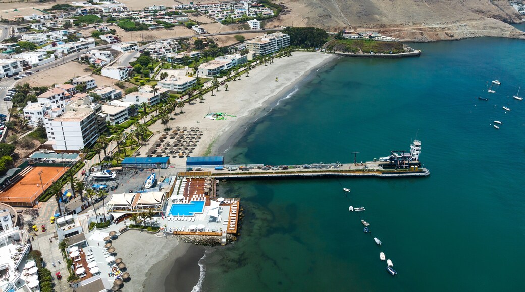 Aerial drone panoramic view of the bay of Ancón in Lima, Peru, showing numerous colorful fishing boats on calm waters with the coastal town and sandy hills in the background.