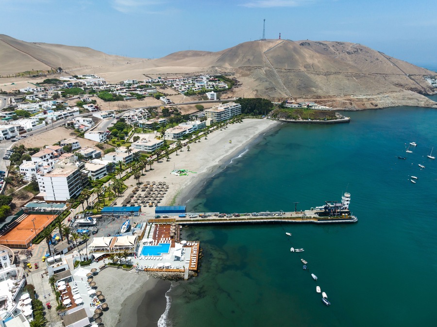 Aerial drone panoramic view of the bay of Ancón in Lima, Peru, showing numerous colorful fishing boats on calm waters with the coastal town and sandy hills in the background.