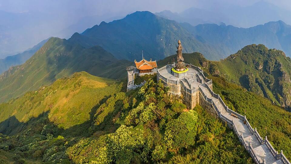 A temple complex on a mountain with a winding path, surrounded by lush greenery and distant mountains.