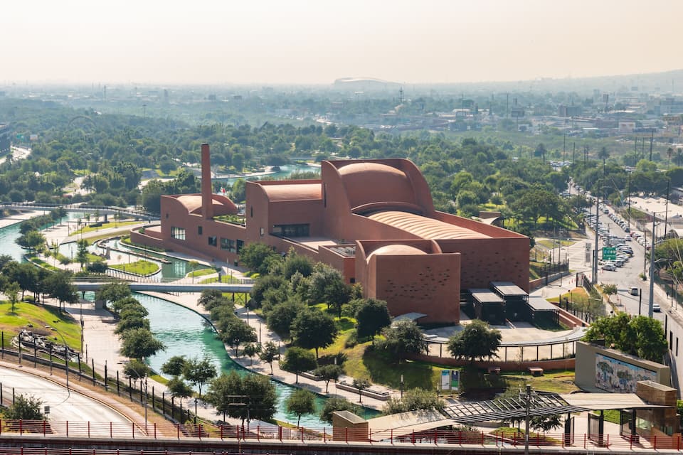 A large, modern building with a curved roof and a prominent chimney, surrounded by a river and greenery.