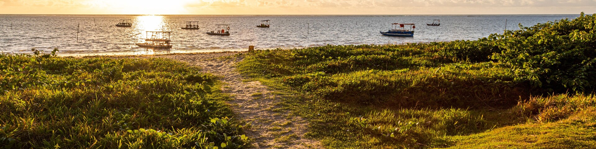 A coastal path leading to the sea with boats and a sunset.