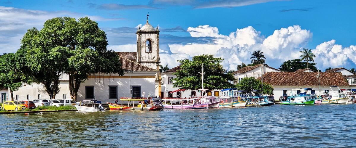 A waterfront with colorful boats, a church with a bell tower, and lush greenery.