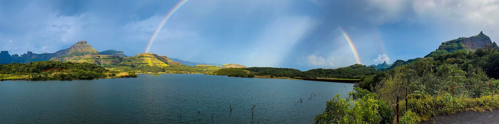 Panoramic view of Ghatghar Dam in Bhandardara featuring a vibrant rainbow over lush green mountains and a serene reservoir after rainfall.