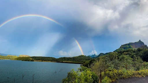 Panoramic view of Ghatghar Dam in Bhandardara featuring a vibrant rainbow over lush green mountains and a serene reservoir after rainfall.