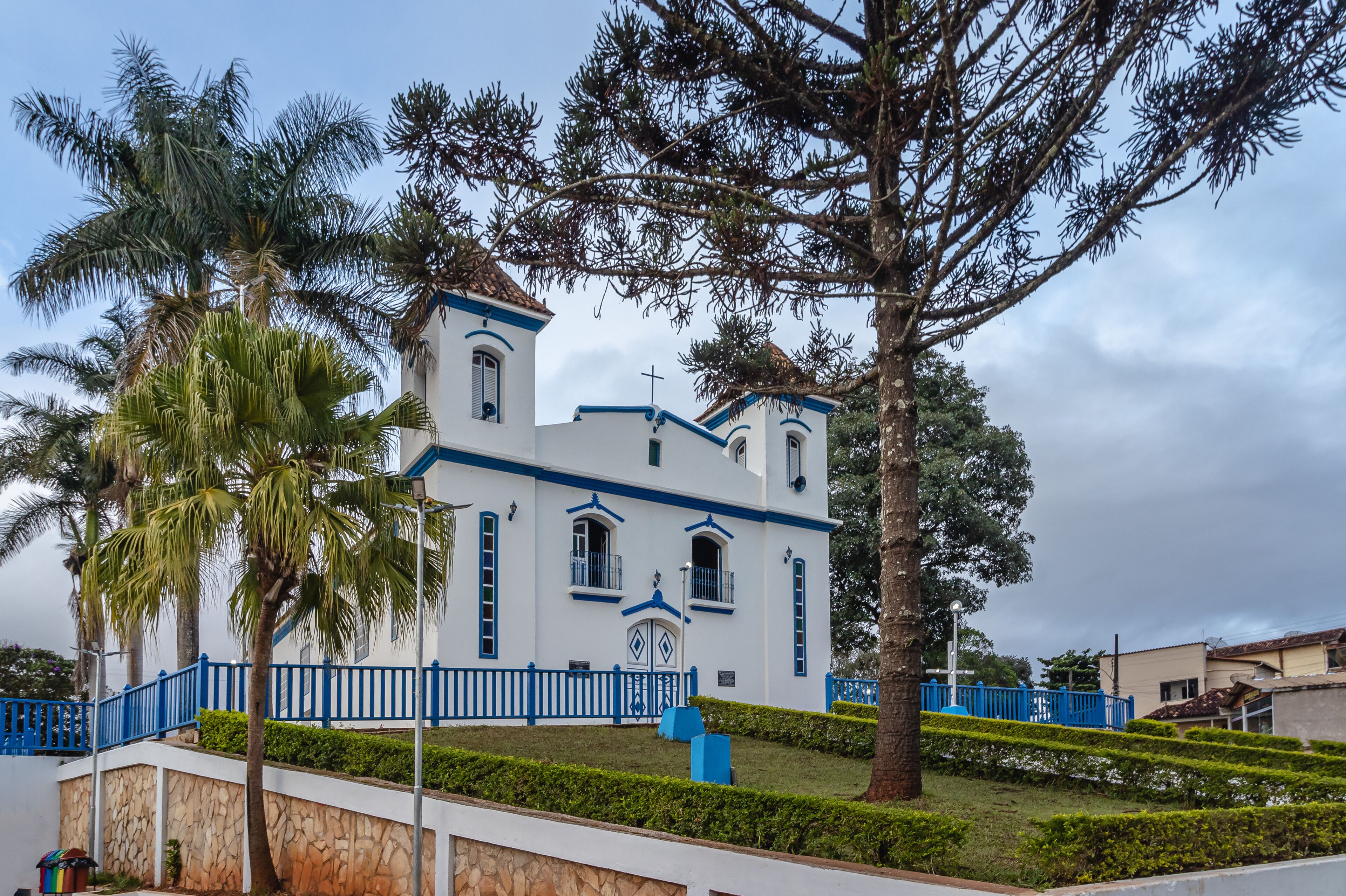 A white church with blue trim and a cross on top, surrounded by palm trees and a blue fence.