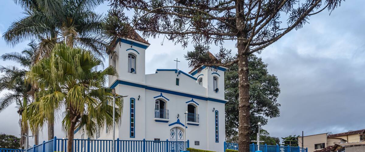 A white church with blue trim and a cross on top, surrounded by palm trees and a blue fence.