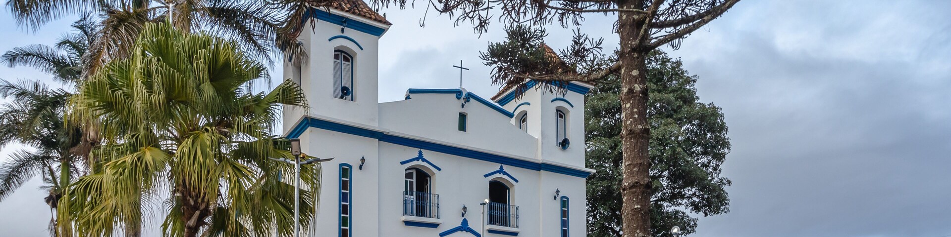 A white church with blue trim and a cross on top, surrounded by palm trees and a blue fence.