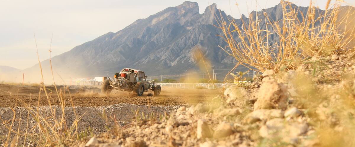 A rugged off-road vehicle in a dry, rocky terrain with sparse vegetation and a mountain range in the background.