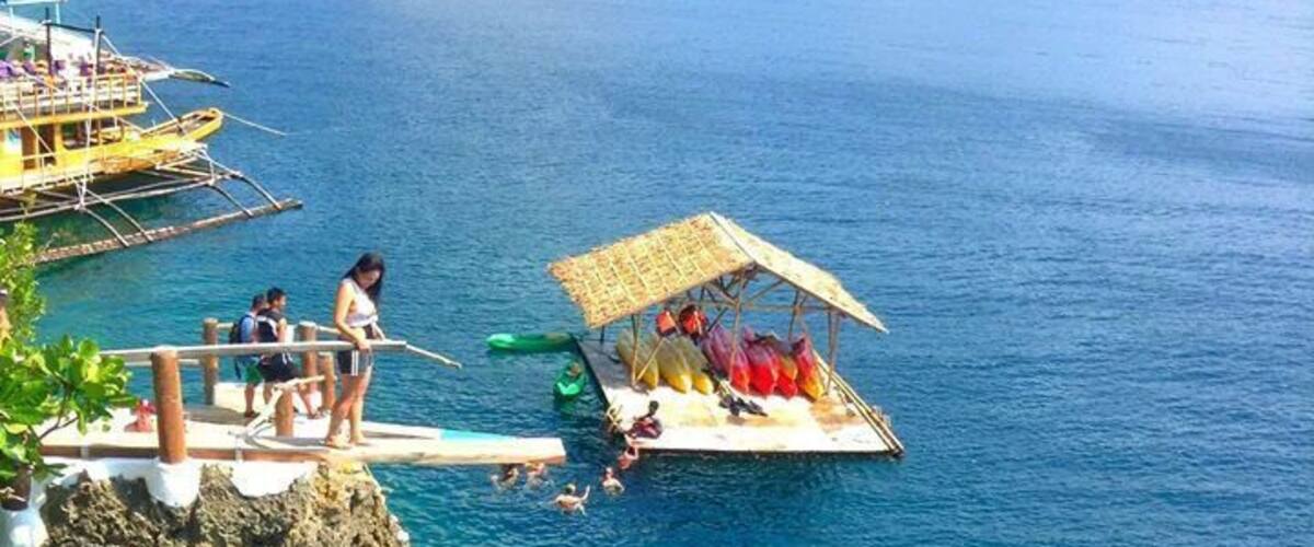 A cliffside with a dock extending into the water, people on the dock, and a small boat with a thatched roof.
