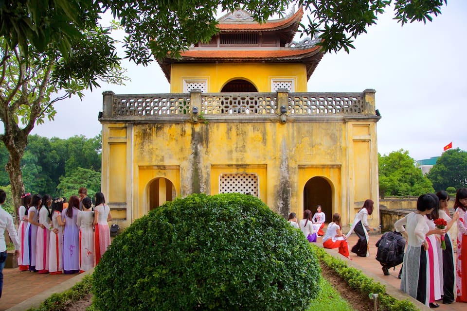 Um edifício amarelo tradicional com telhado de azulejos e decorações ornamentadas, rodeado por vegetação exuberante e um grupo de pessoas vestidas com trajes típicos.