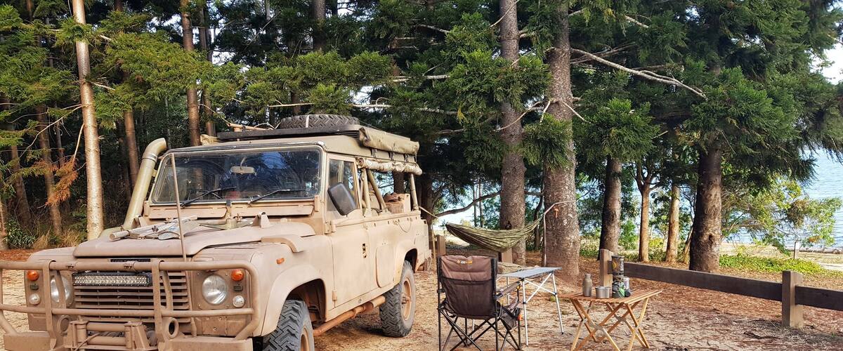 A beige off-road vehicle parked in a forested area with a hammock and camping gear.
