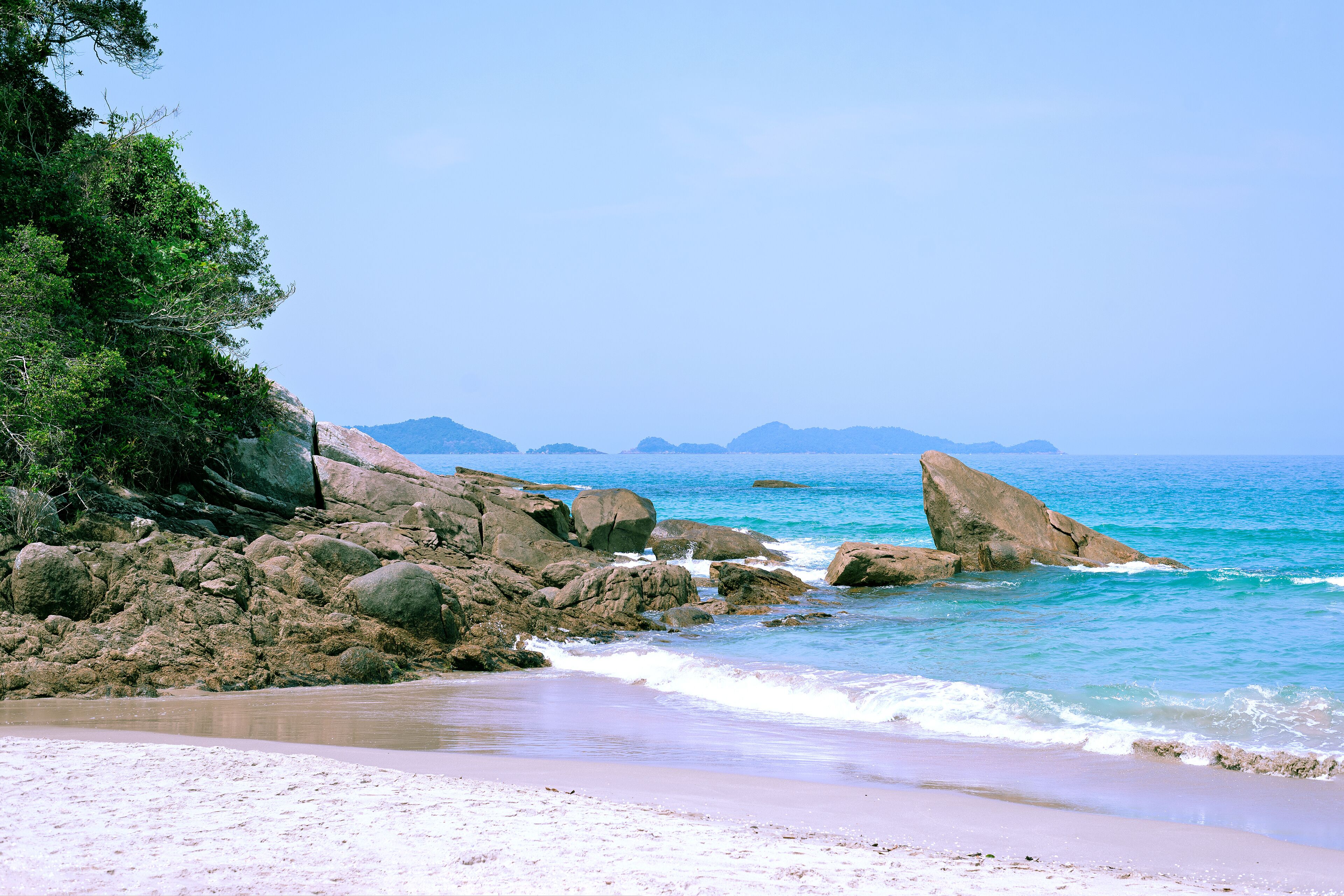 A beach with large rocks, clear blue water, and distant mountains.