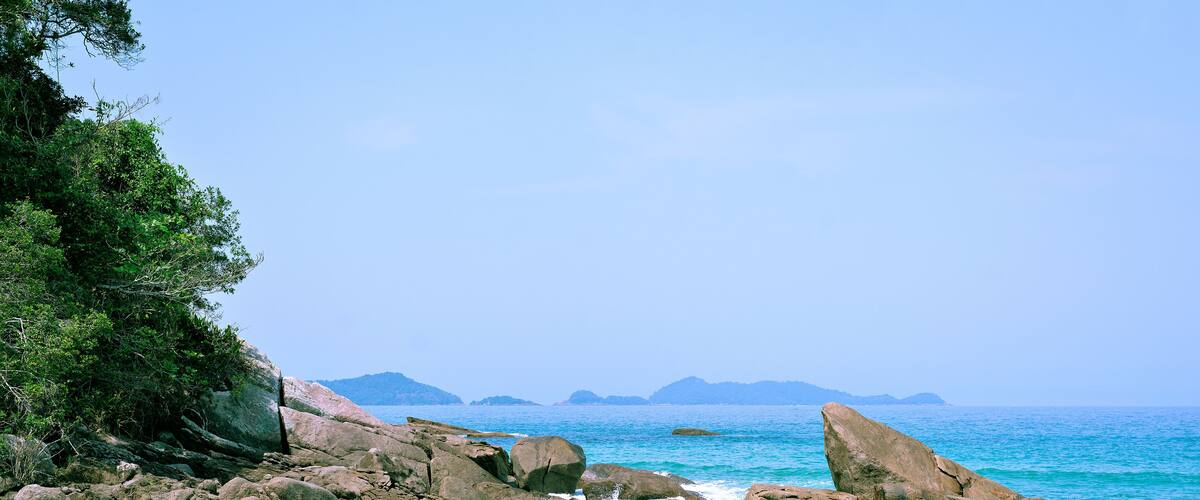 A beach with large rocks, clear blue water, and distant mountains.