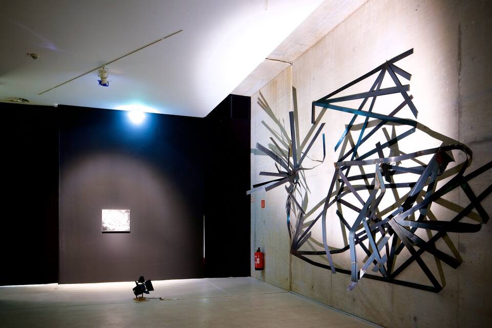 Een kunstgalerie met een grote abstracte metalen sculptuur aan de muur en een kleine schotelantenne op de voorgrond.