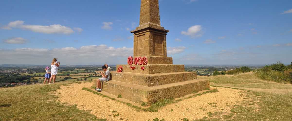 A tall stone monument with red flowers on its base, surrounded by grassy terrain and a clear blue sky.