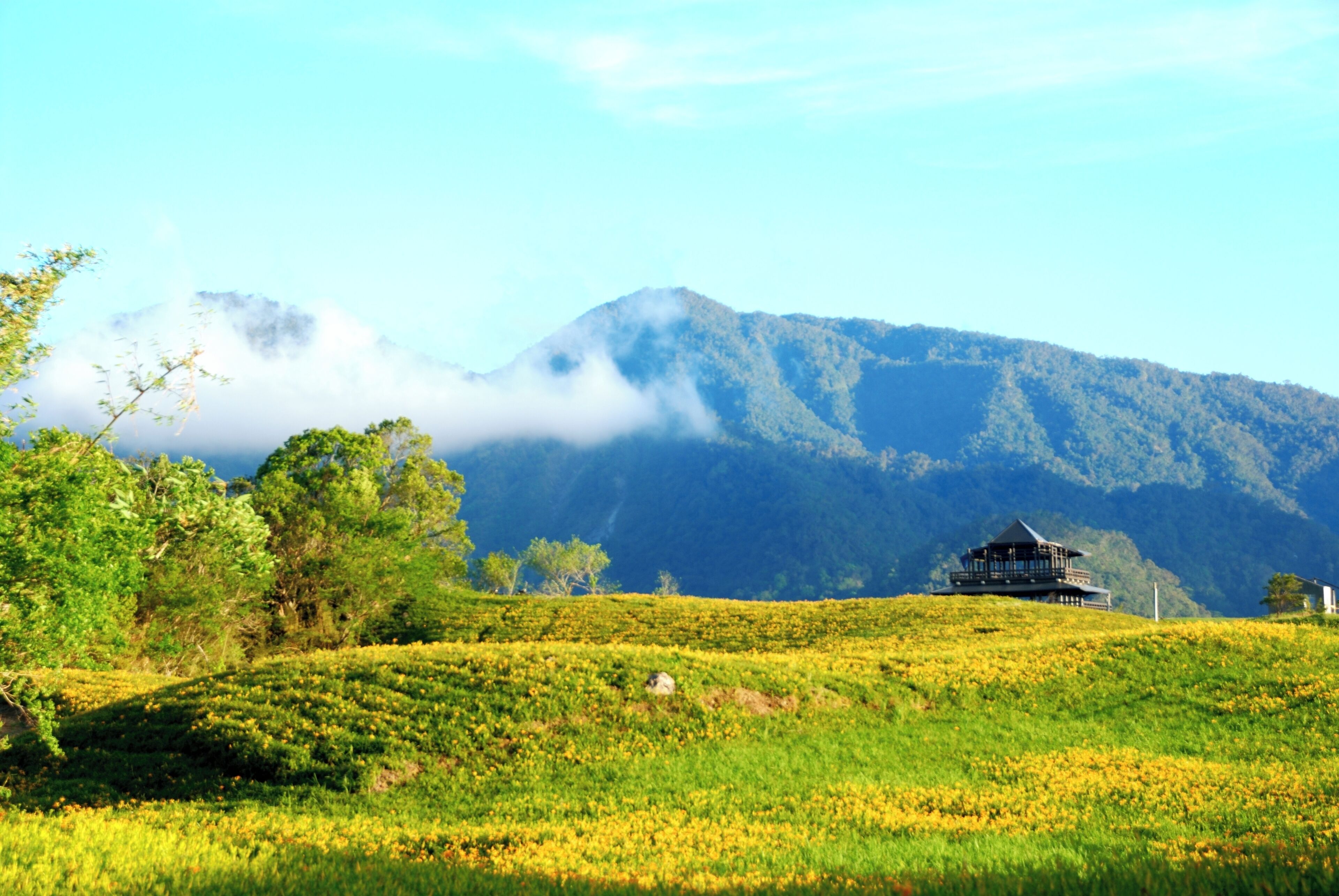 A lush green landscape with a traditional building on a hill, surrounded by mountains and clouds.