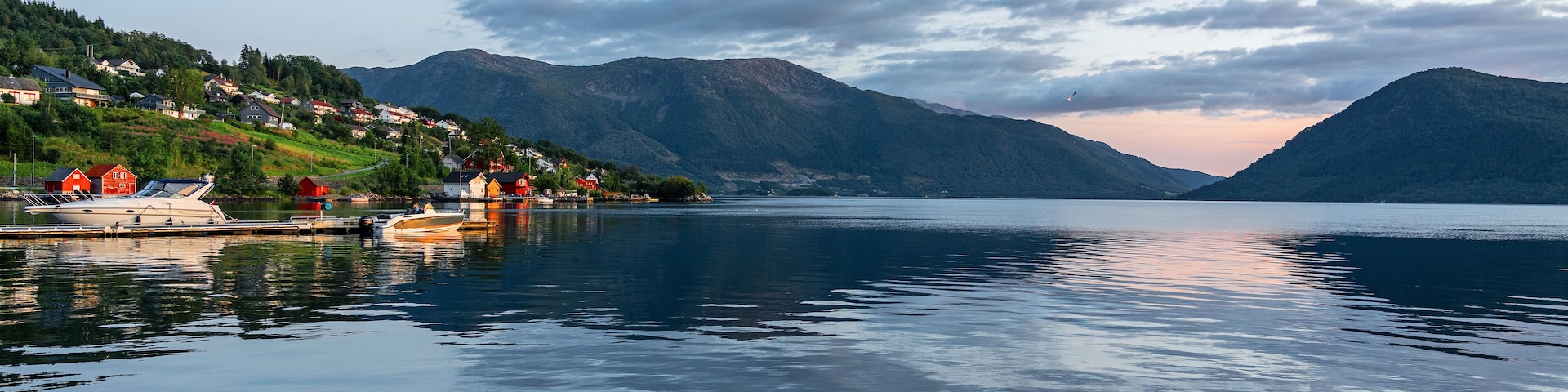 Um lago calmo com barcos atracados à esquerda, rodeado por casas e montanhas ao fundo.