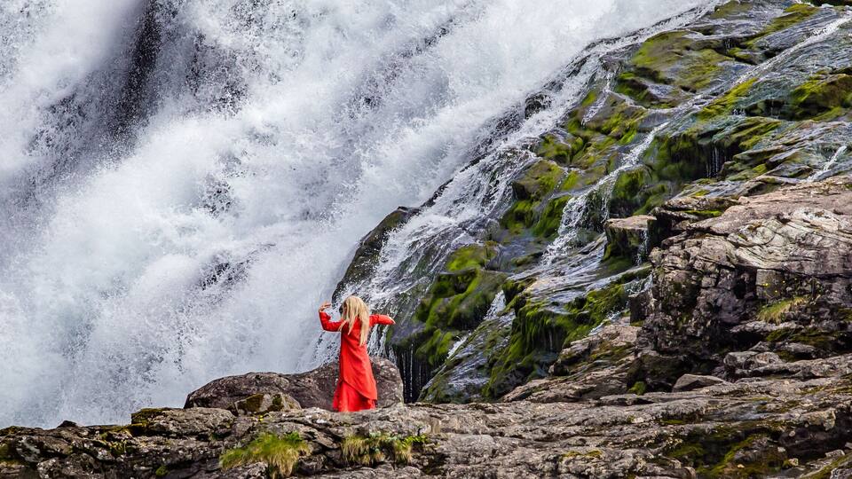 A waterfall cascading down a rocky cliff with a person in a red dress standing on the rocks.
