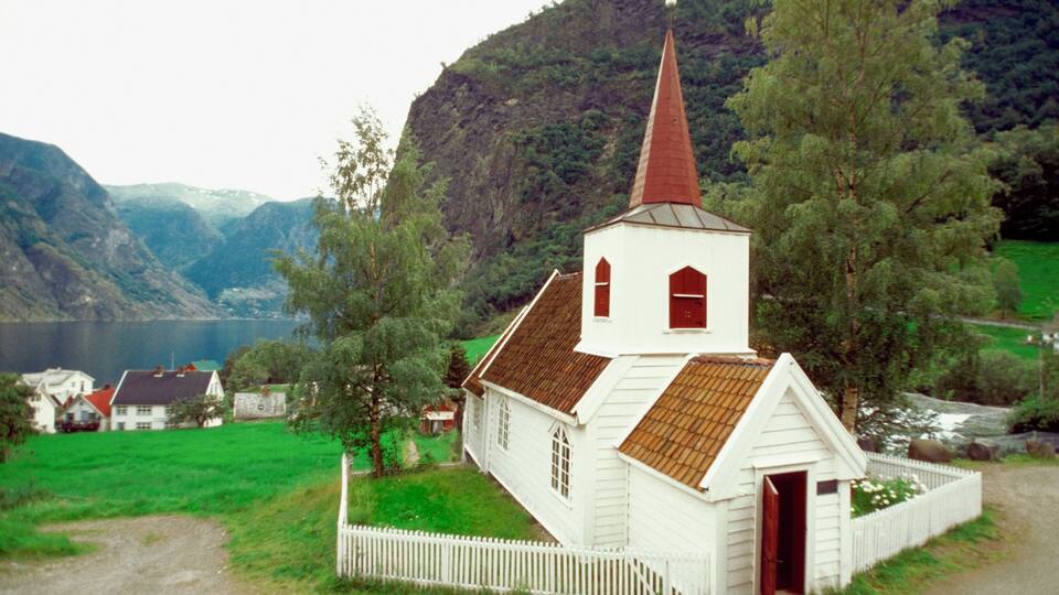 Una iglesia blanca con campanario rojo, rodeada de campos verdes y montañas.