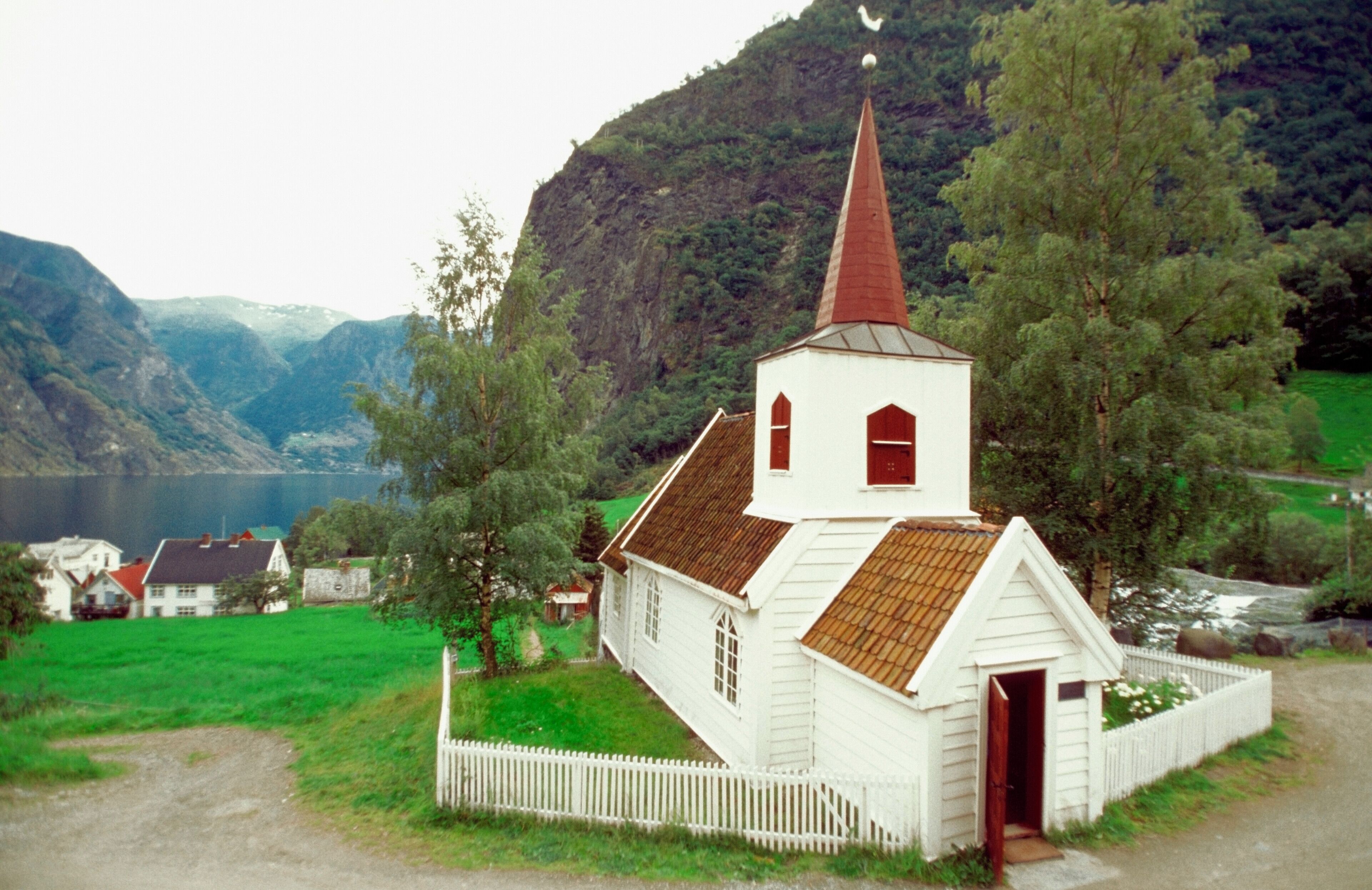 A white church with a red steeple surrounded by green fields and mountains.