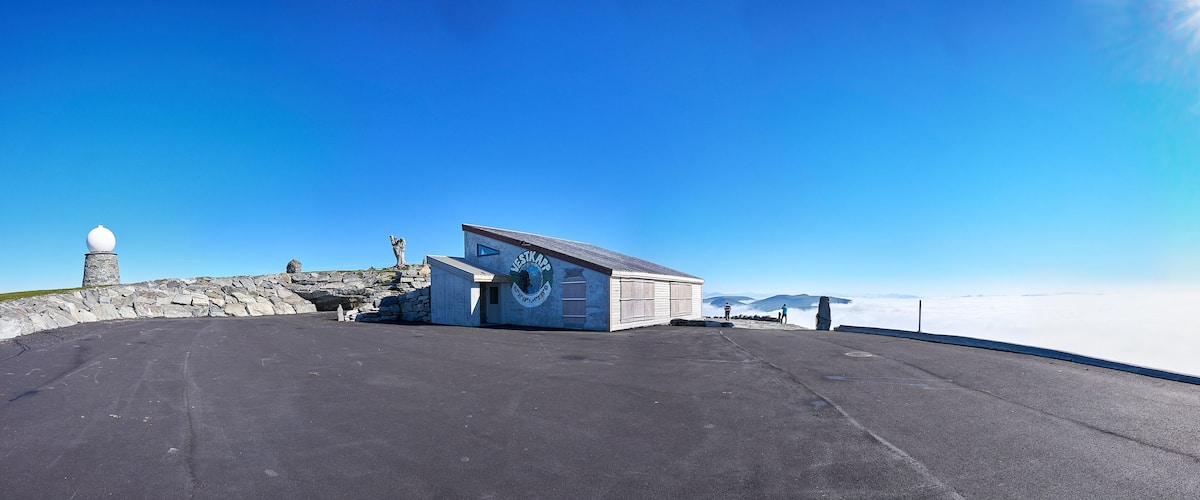 A clear sky, a stone monument, a wooden building, and a paved area with a view of mountains.