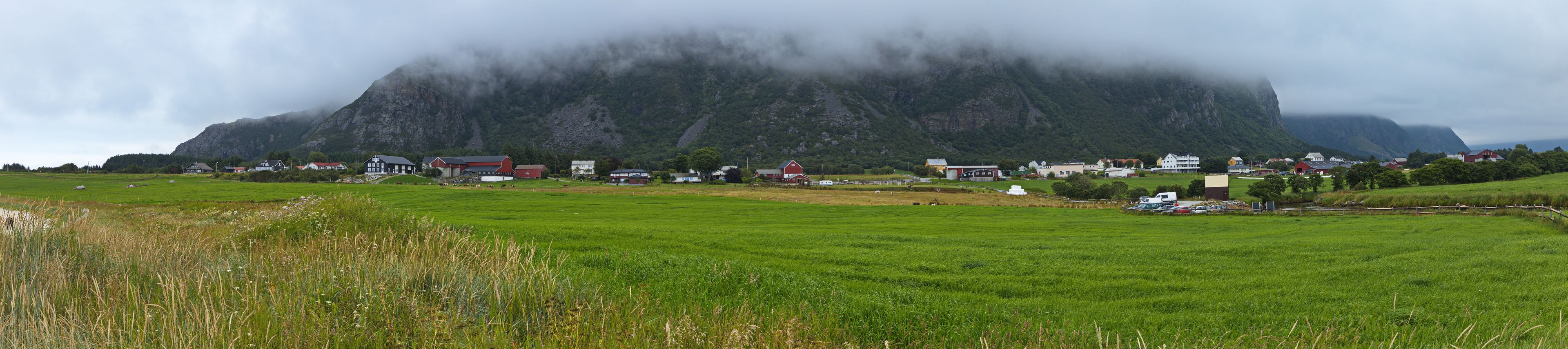 A rural landscape with a prominent mountain in the background, a small village with houses, and a green field in the foreground.