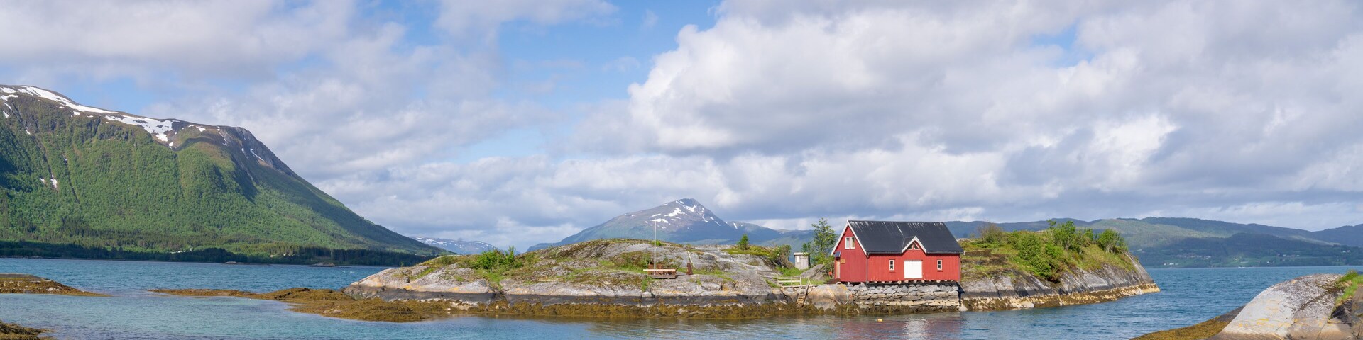 Una casa roja en una pequeña isla rodeada de agua, con montañas al fondo.