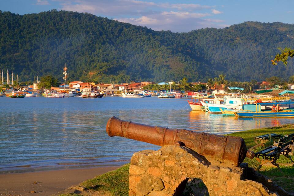 Uma vila costeira com barcos, um canhão e montanhas ao fundo.