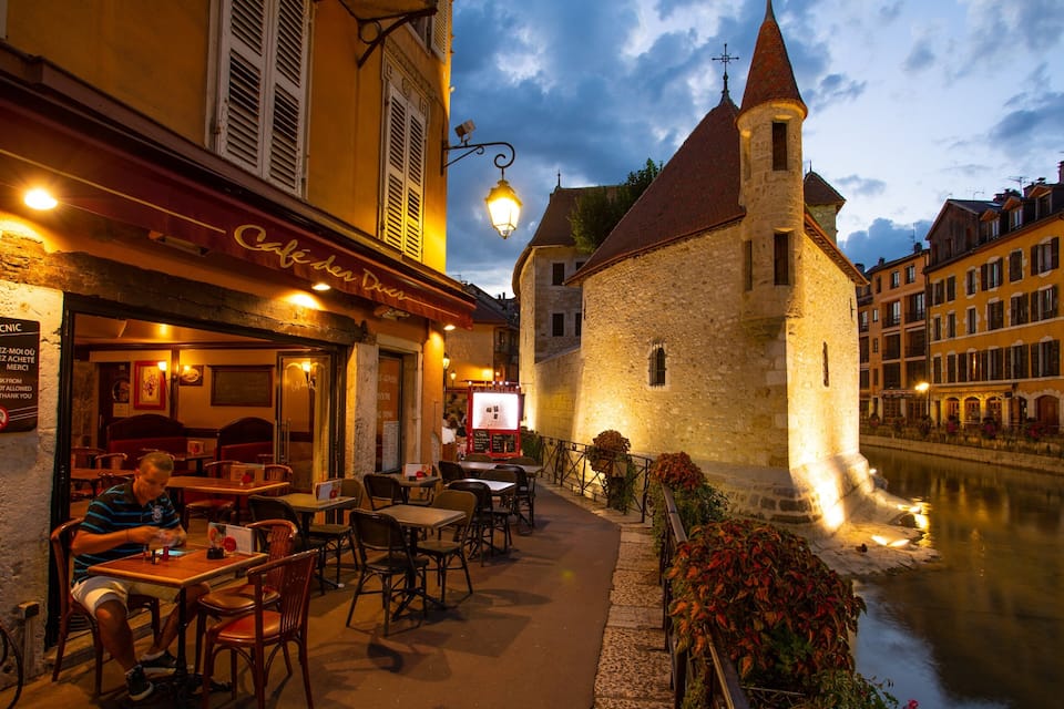 A riverside cafe with outdoor seating, a stone tower, and historic buildings.