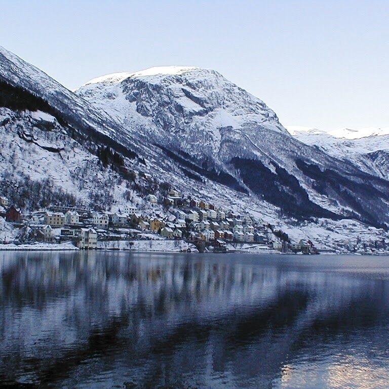 A snow-covered mountain range with a town at the base, reflecting in a calm lake.