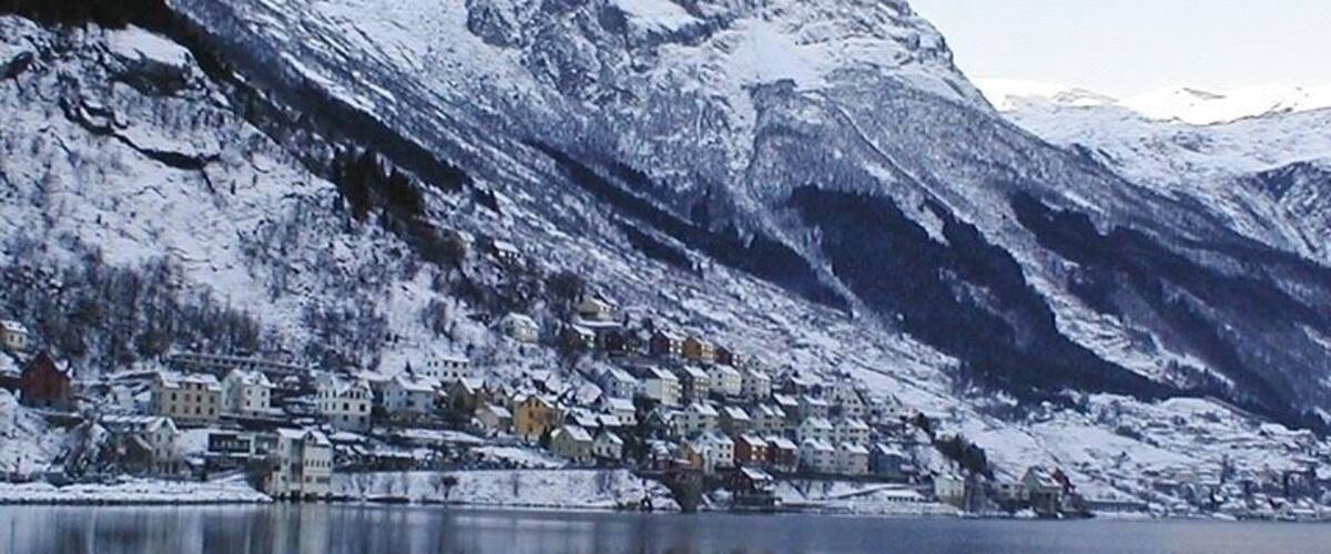 A snow-covered mountain range with a town at the base, reflecting in a calm lake.