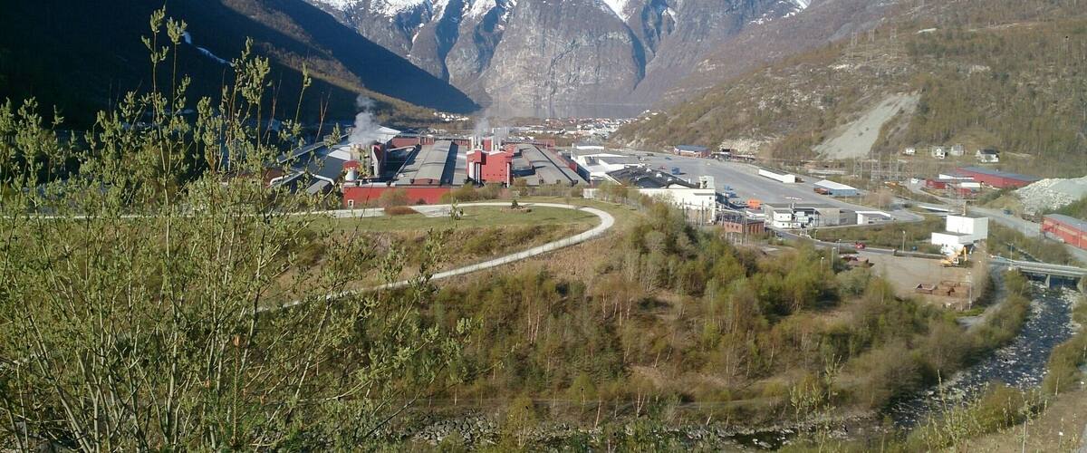 A mountainous landscape with a snow-capped peak in the background, a river flowing through the valley, and a large industrial complex with red buildings.