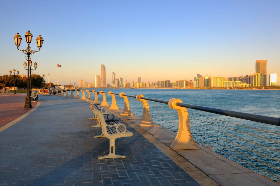 A waterfront promenade with benches and lamp posts, overlooking a city skyline with high-rise buildings.