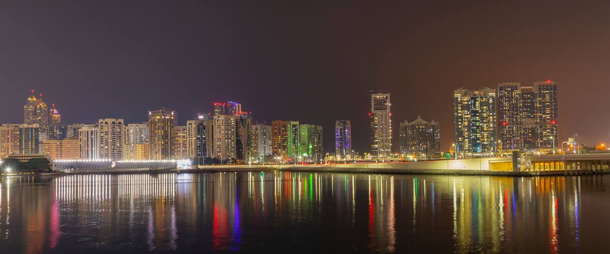 A nighttime city skyline with illuminated buildings reflected in calm waters.
