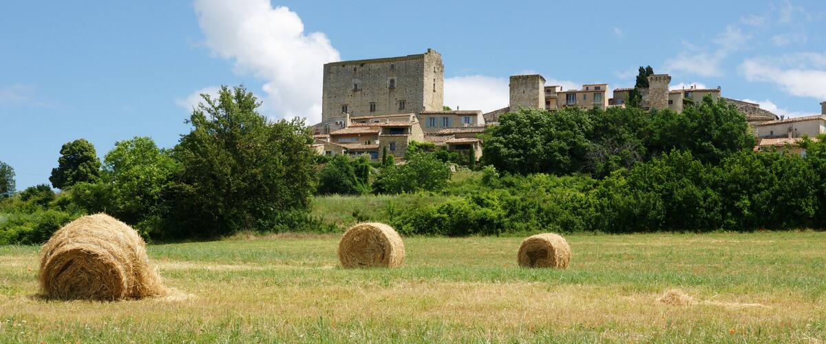 A rural landscape with hay bales in a field, a historic stone building, and lush greenery in the background.