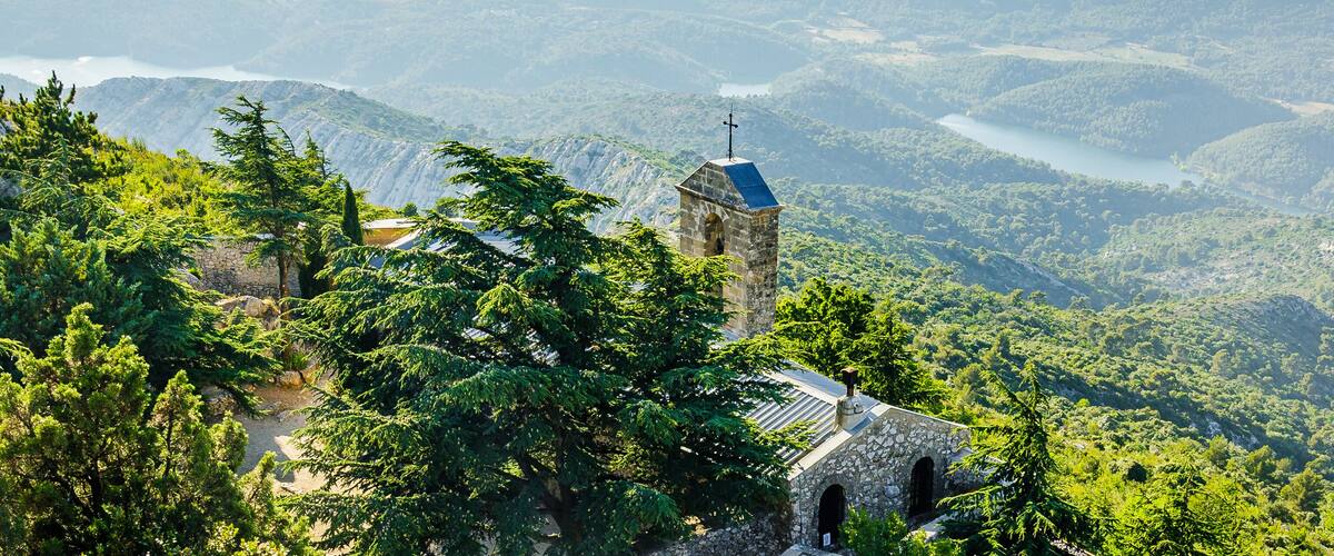 A stone church with a bell tower surrounded by lush green forest and a lake in the background.