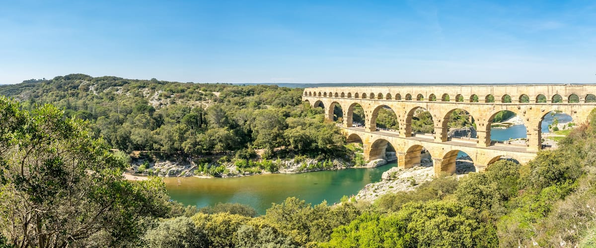 A stone aqueduct over a river, surrounded by dense forest and hills.
