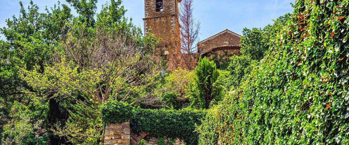 A stone pathway leading to a tower with a bell, surrounded by greenery and a clear sky.