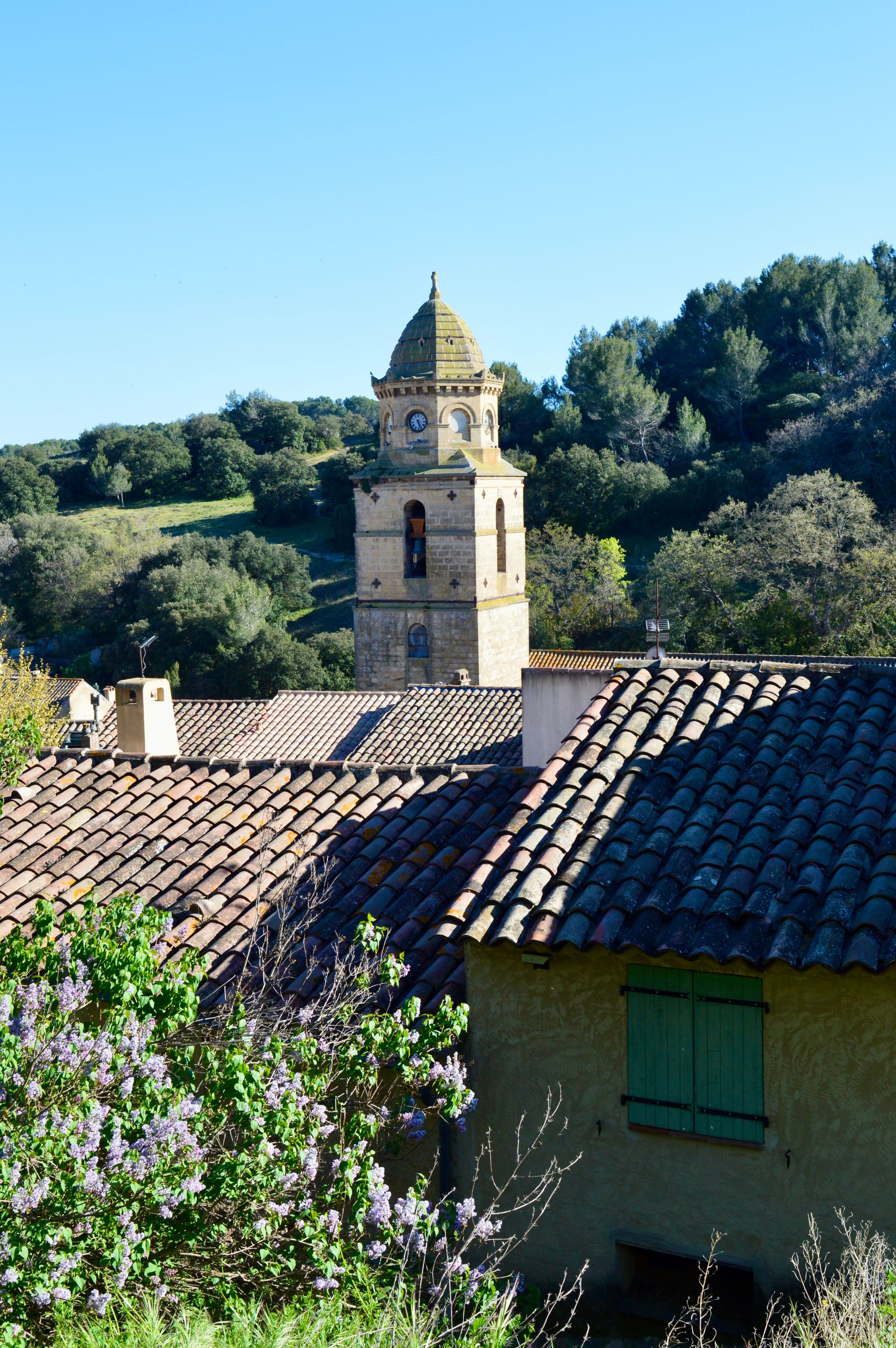 Une tour surmontée d’un dôme et d’une horloge, entourée d’un paysage rural avec des arbres et un toit en tuiles.
