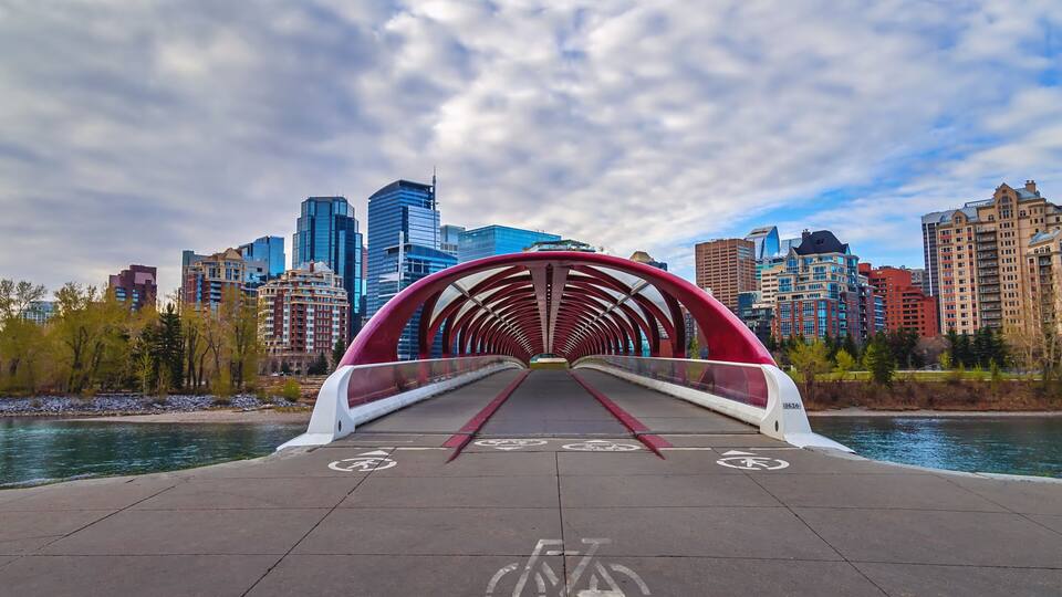 Un puente rojo para bicicletas sobre un río, con el horizonte de una ciudad al fondo.