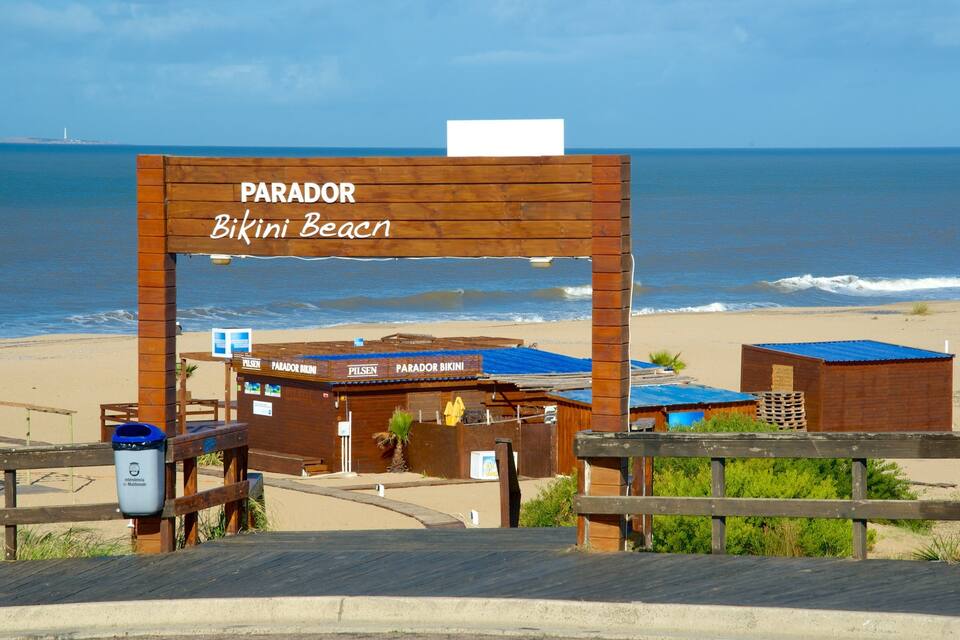 A wooden sign with 'PARADOR Bikini Beach' written on it, overlooking a beach with a wooden fence and a small building with a blue roof.