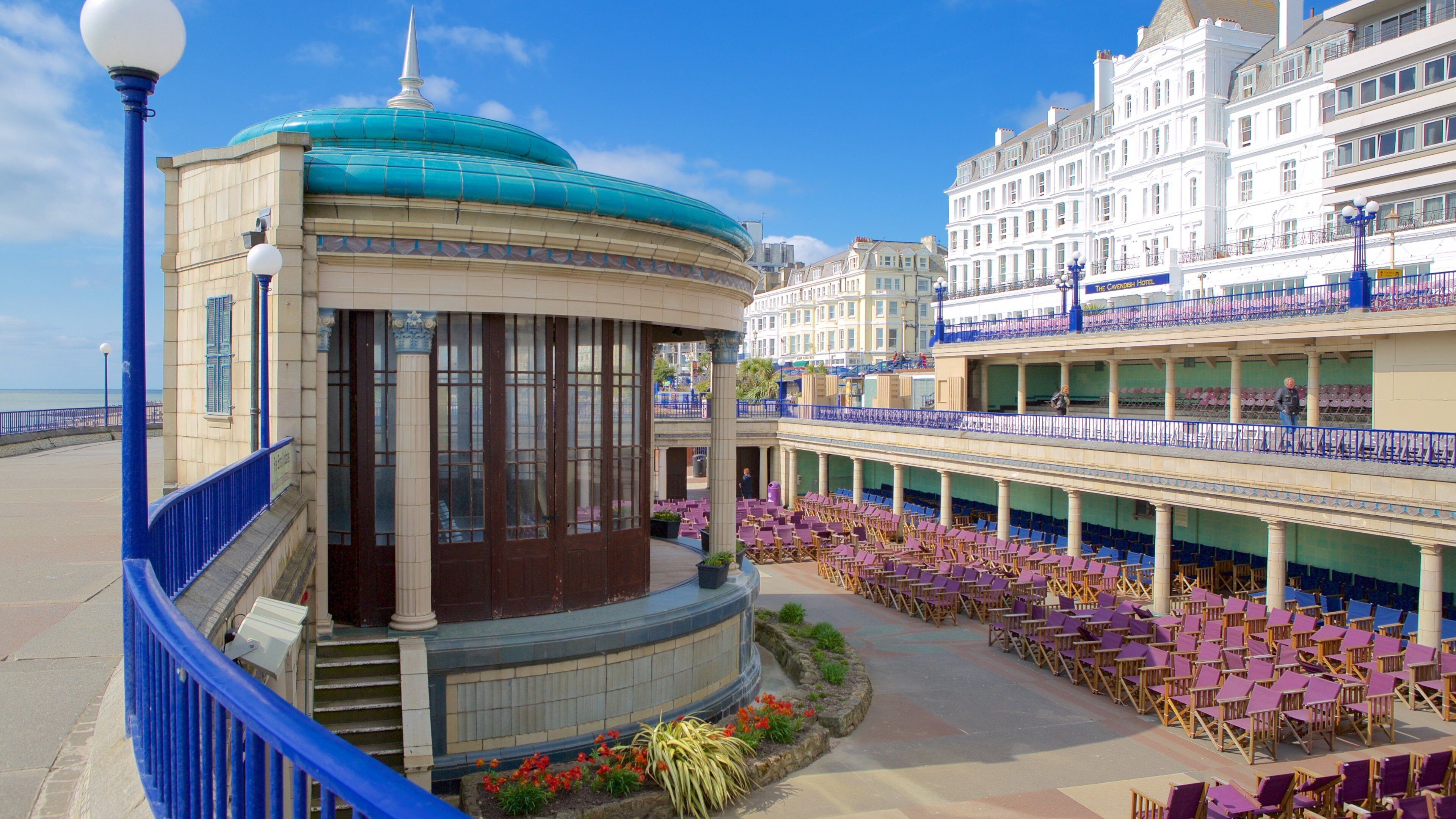 A seaside promenade with a covered seating area, a blue-domed building, and a hotel with balconies.