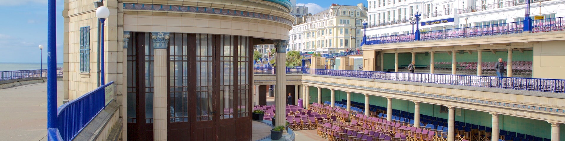 A seaside promenade with a covered seating area, a blue-domed building, and a hotel with balconies.