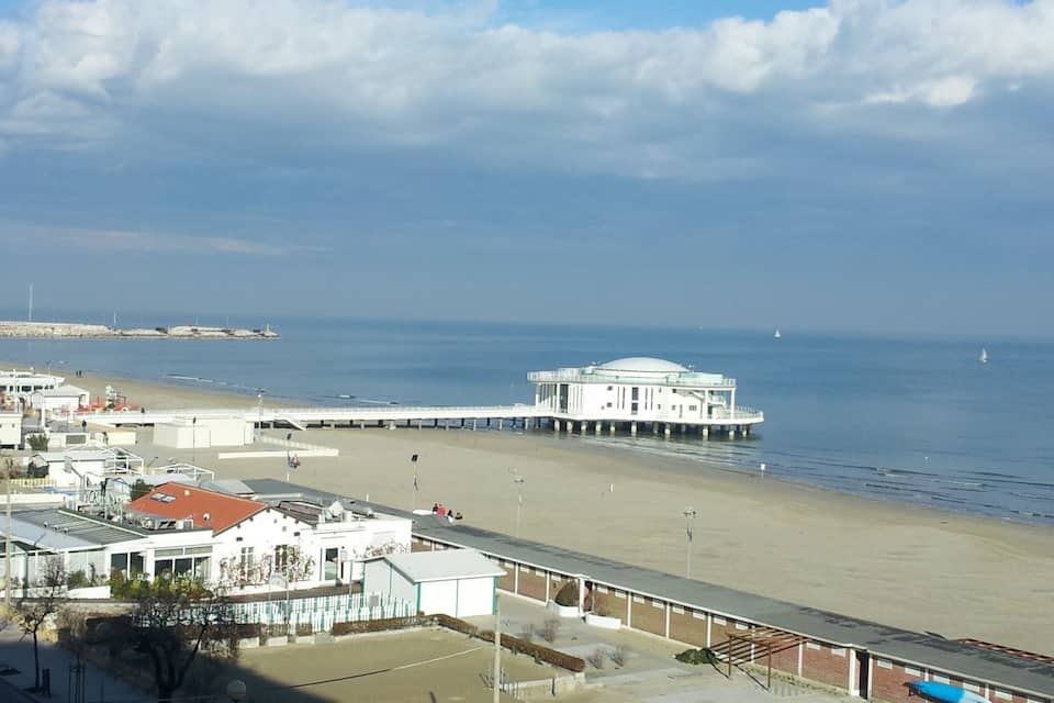 A beach with a pier extending into the sea, a white building with a dome, and a sandy shore with buildings in the background.
