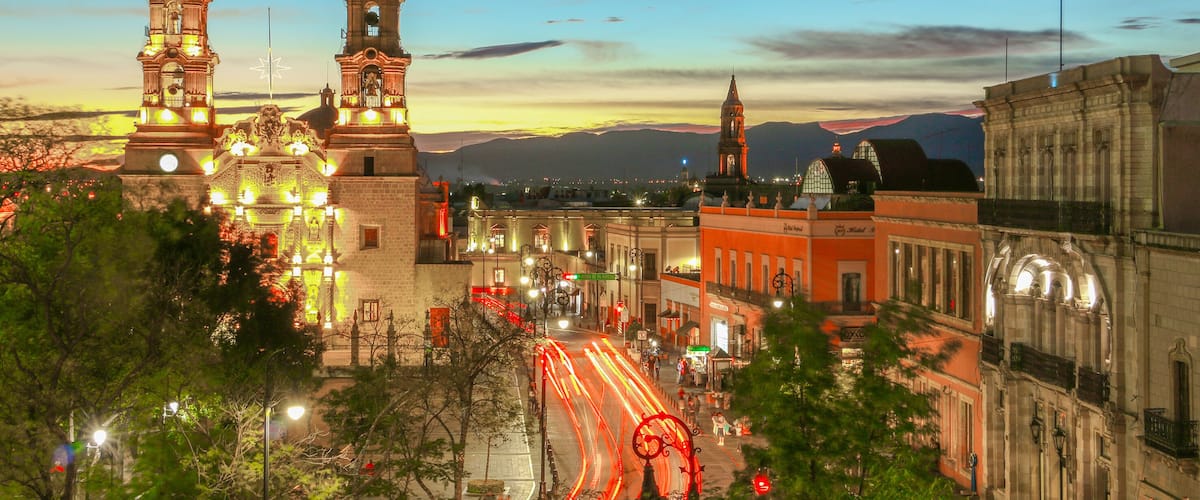 Un paisaje urbano con torres iluminadas, una iglesia destacada y una carretera serpenteante con estelas de luz.