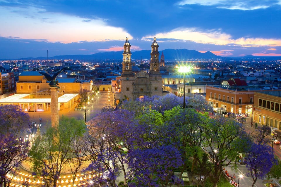 Un paisaje urbano al anochecer con edificios iluminados, una iglesia destacada con dos torres y una cadena montañosa al fondo.