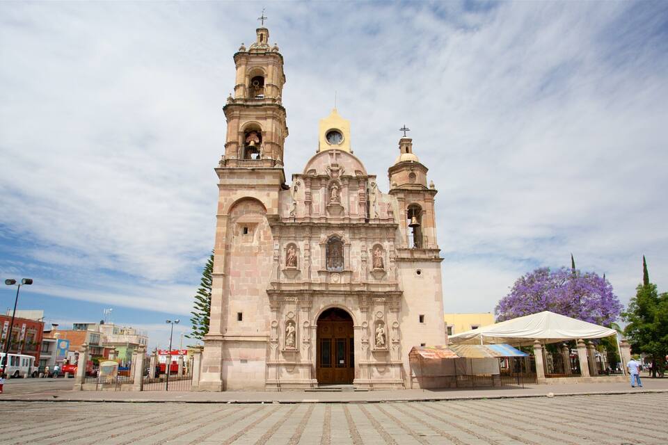 A historic church with two bell towers and a dome, surrounded by a paved plaza and modern buildings.