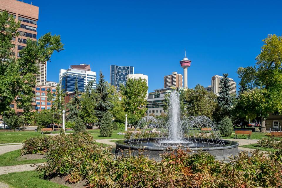 Um parque urbano com uma fonte, rodeado por árvores e edifícios, incluindo uma torre.