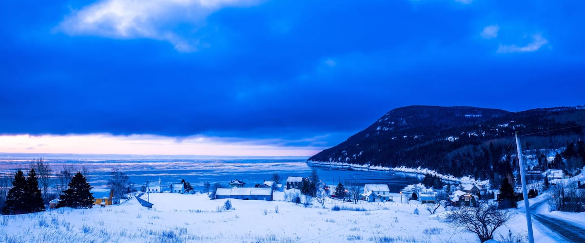 A snowy landscape with a river, houses, and mountains under a blue sky.